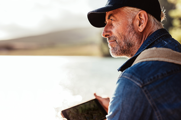 Older male holding a hand-held electronic while looking thoughtfully over a lake. 